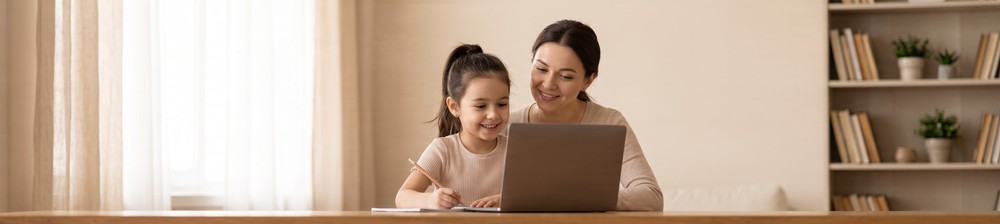 Mother and young daughter smiling at a laptop during an online Russian lesson